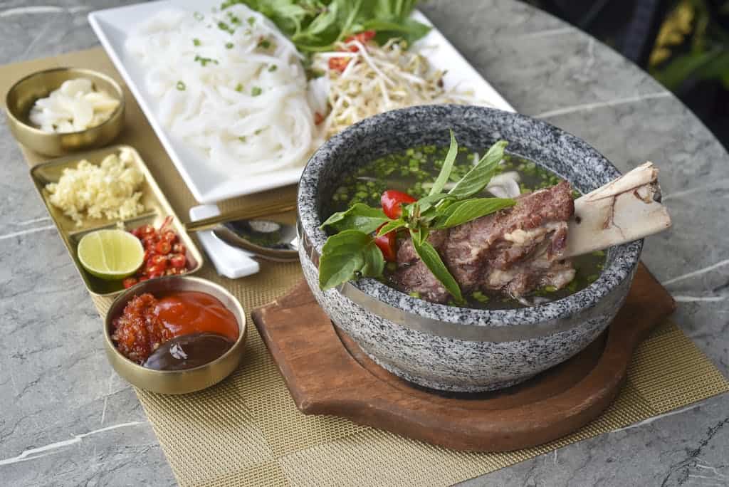 A delicious bowl of Vietnamese pho with beef bones and fresh garnishes served on a stone table.