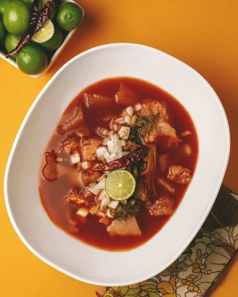 A delicious Mexican menudo soup, honeycomb tripe, garnished with lime and onions served on a vibrant background.