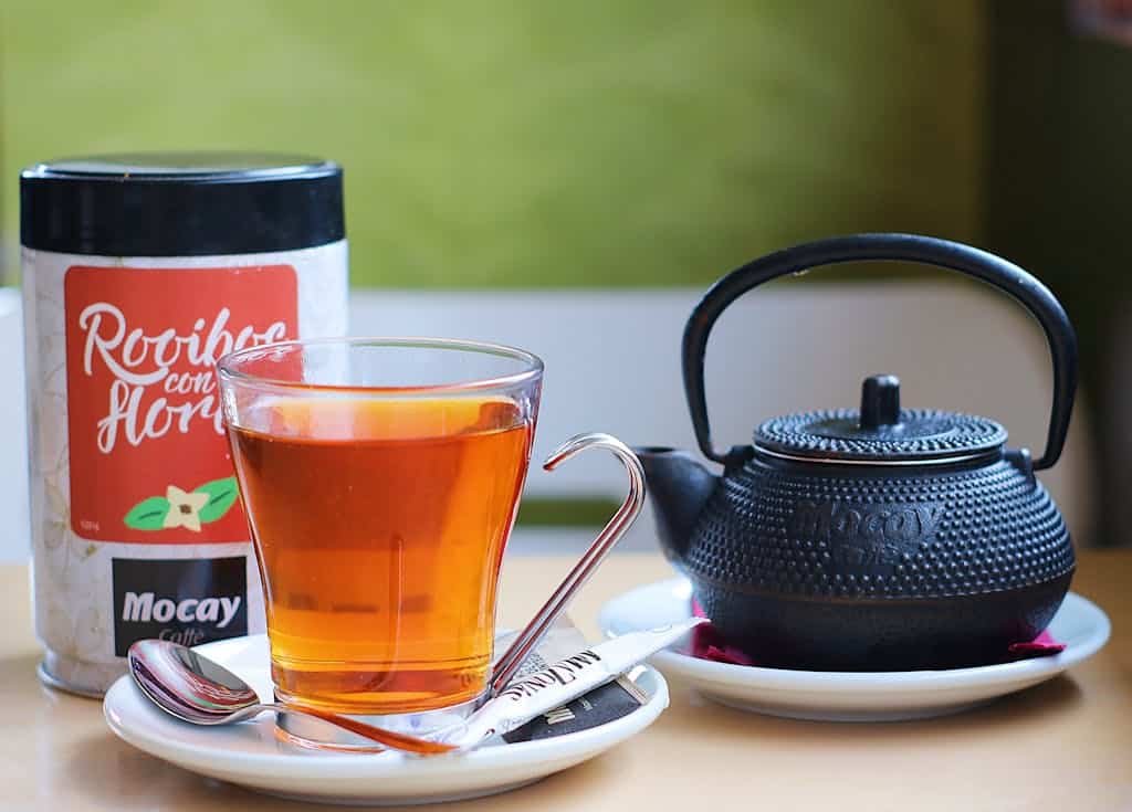 A stylish black teapot paired with a clear glass of rooibos tea, showcasing a cozy tea setup indoors.