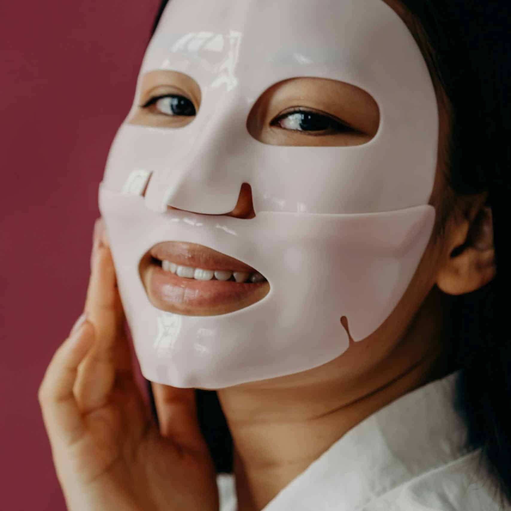 Beautiful Asian woman enjoying skincare routine with a hydrating sheet mask against a red background.