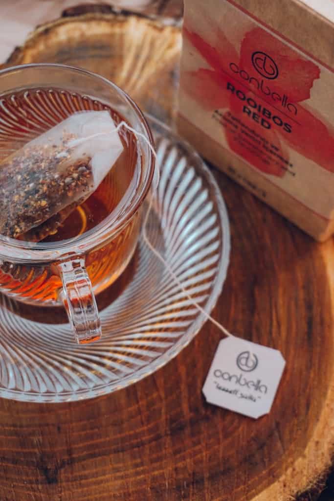 Close-up of a glass cup with rooibos tea and a teabag on a wooden surface.