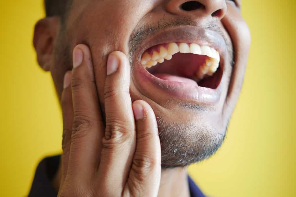 Close-up of a man holding his cheek, displaying a wide open mouth with healthy teeth against a yellow background.