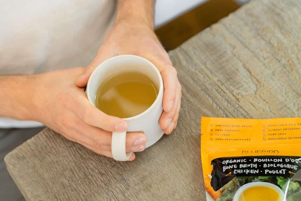 Close-up of hands holding a cup of bone broth beside a package on rustic table.