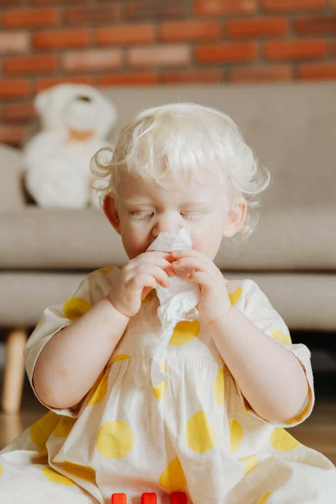 Cute blonde child in a yellow polka dot dress sneezing indoors with a tissue. Bright and warm atmosphere.