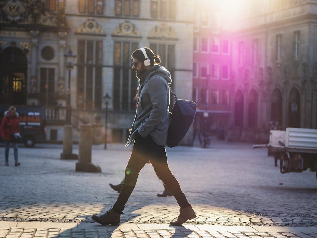 Man with headphones walks down a sunlit urban street, casting a shadow on the cobblestones.